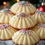 A plate of beautifully piped Christmas whipped shortbread cookies, decorated with festive red and green sprinkles and a dusting of powdered sugar, set against a cozy holiday background with warm lights and ornaments.