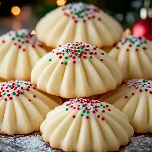 A plate of beautifully piped Christmas whipped shortbread cookies, decorated with festive red and green sprinkles and a dusting of powdered sugar, set against a cozy holiday background with warm lights and ornaments.